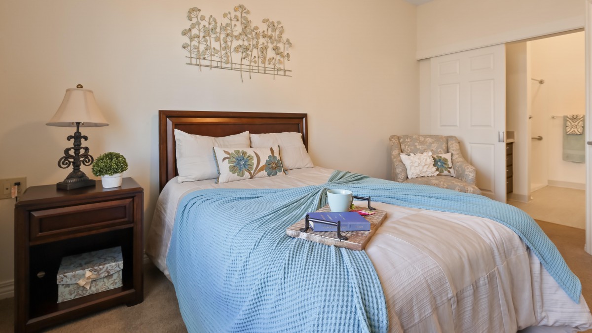 A cozy bedroom with a bed adorned with pillows and a blue blanket. A tray on the bed holds a book and a mug.
