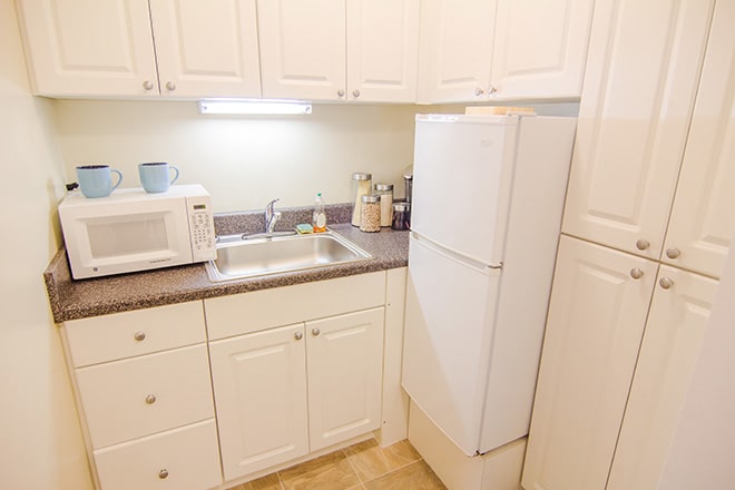 A compact kitchen with white cabinets, featuring a microwave, refrigerator, and sink.