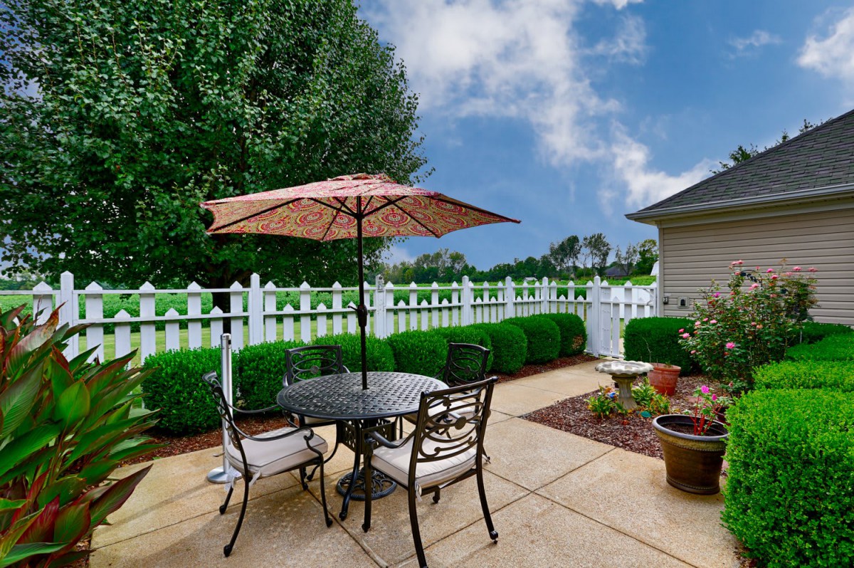 A cozy backyard patio with a table, chairs, and an umbrella next to a lush garden and white picket fence.