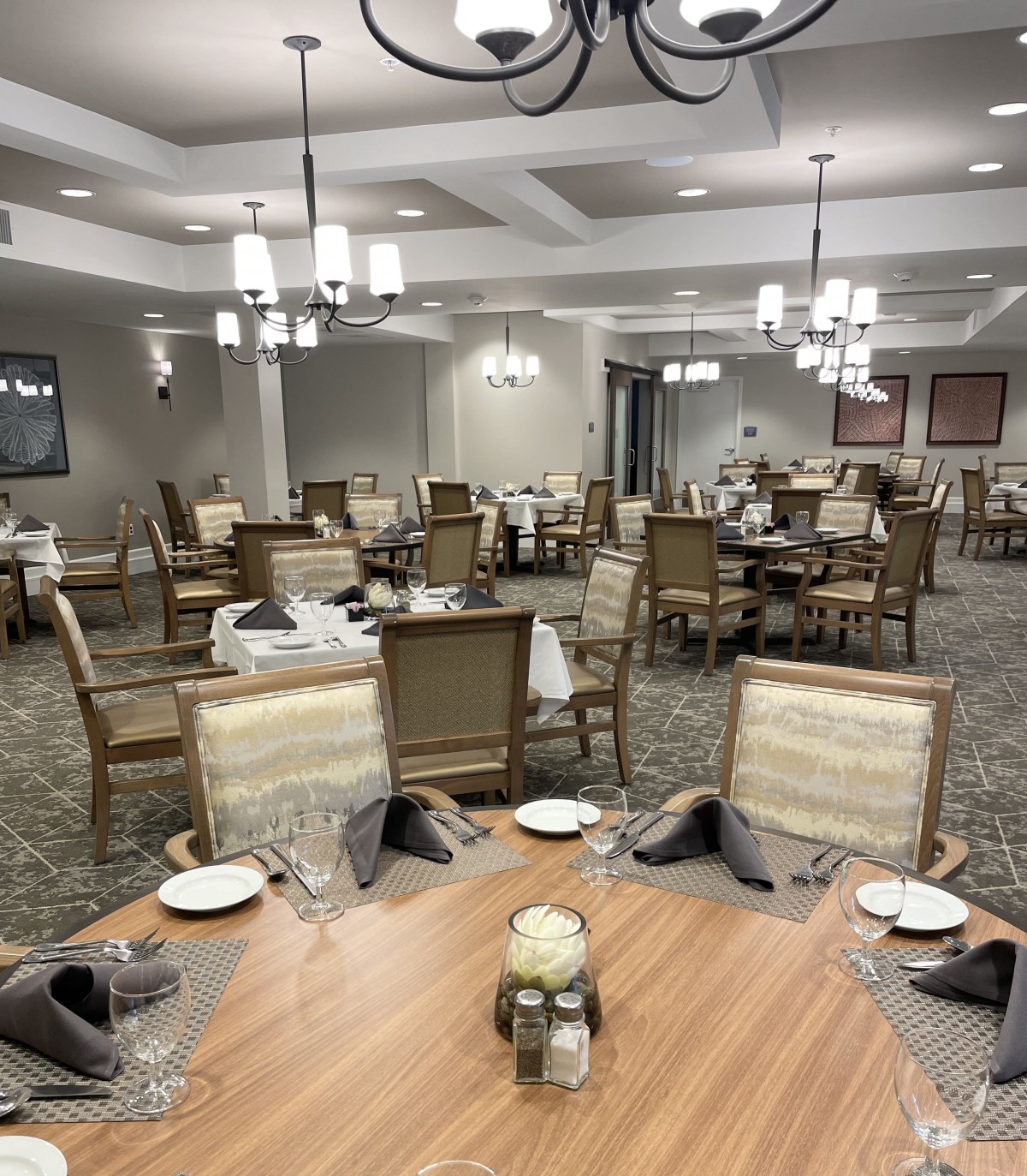 An elegantly set dining room with multiple tables and chairs, decorated with chandeliers and place settings.