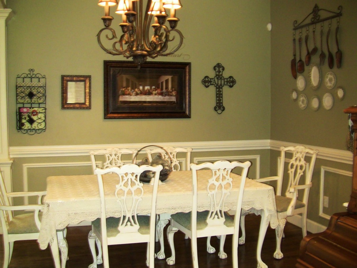 A formal dining room with a chandelier, ornate white table and chairs, decorated with wall art and a cross on green walls.