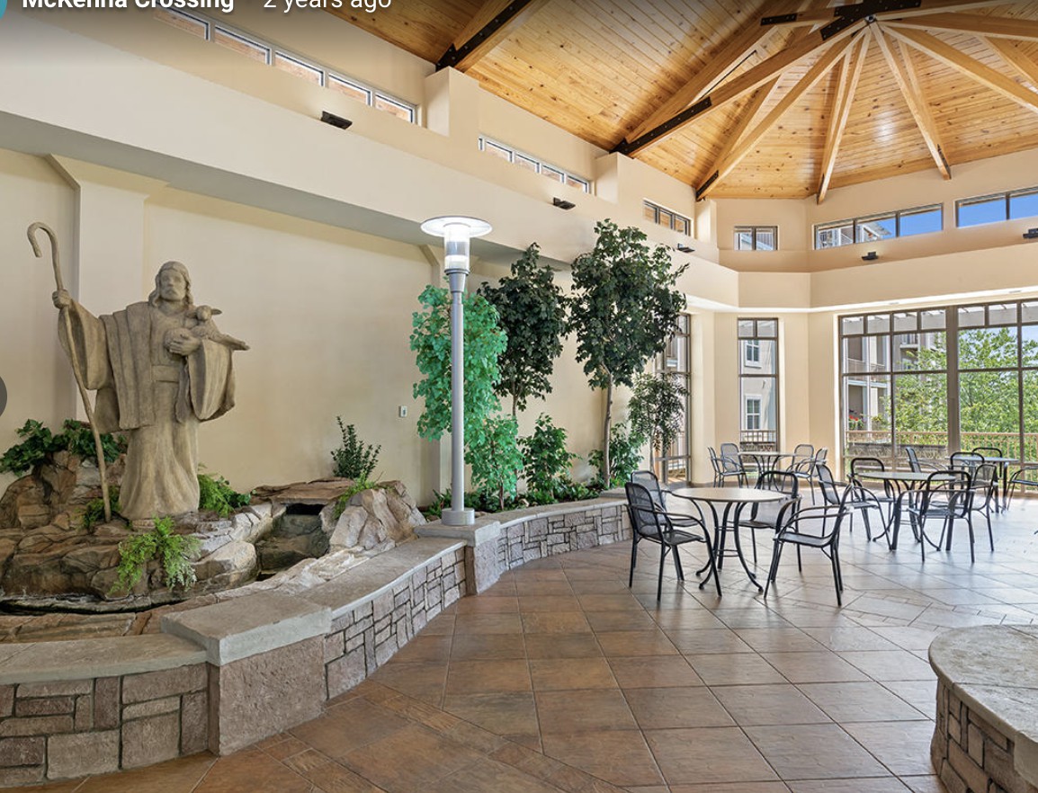 A serene indoor space featuring a statue and fountain, surrounded by greenery and a seating area under an arched wooden ceiling.