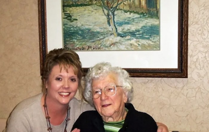 A younger woman and an elderly woman smiling together indoors with a painting in the background.