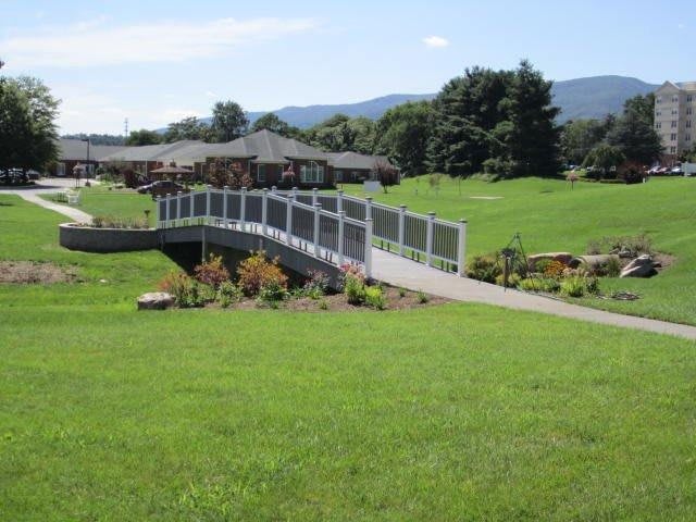 A scenic view of a garden with a bridge leading to a building, surrounded by lush greenery and hills in the background.