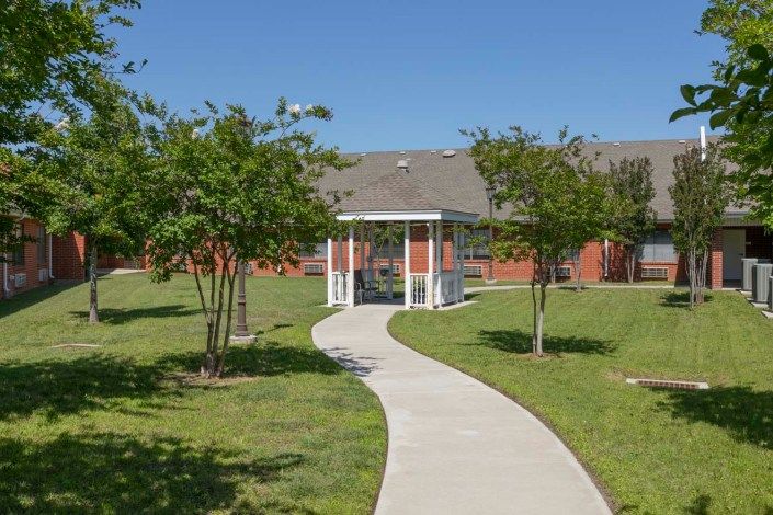 A paved pathway leads to a white gazebo surrounded by green lawns and trees, with a building in the background.