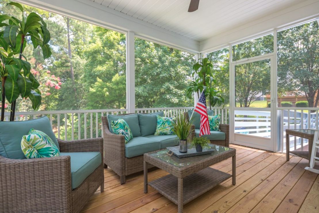 A cozy screened porch with wicker furniture, cushions, and a potted plant, overlooking lush greenery.