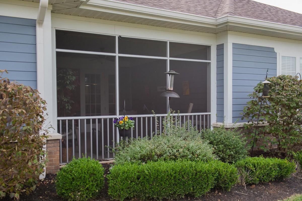 A screened porch with a garden view, featuring shrubs and a bird feeder.