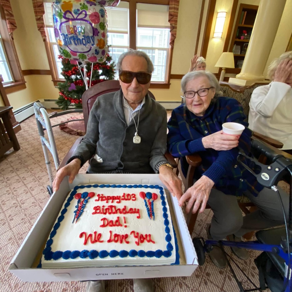 Elderly individuals celebrating a birthday with a large cake and balloons.