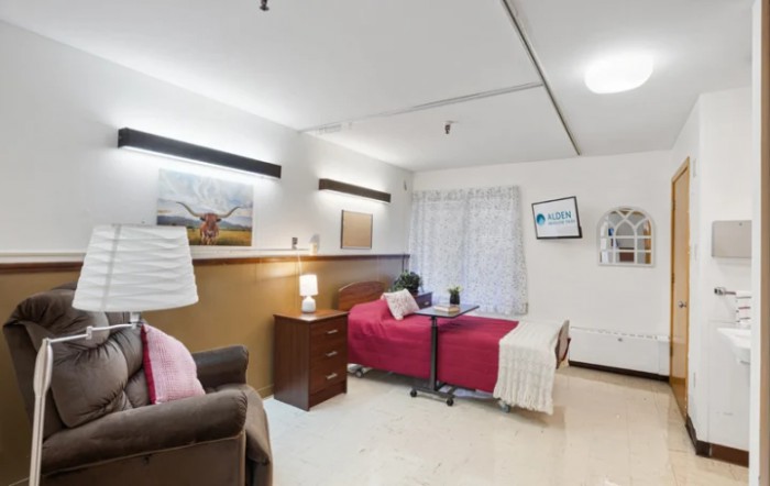 A cozy living space with a comfortable armchair, bedside table with a lamp, and a bed adorned with pink covers.
