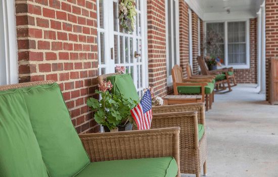 A row of wicker chairs with green cushions on a brick porch, featuring potted plants and small American flags.
