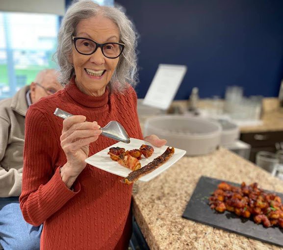 A smiling woman with glasses holding a plate of food in a kitchen.