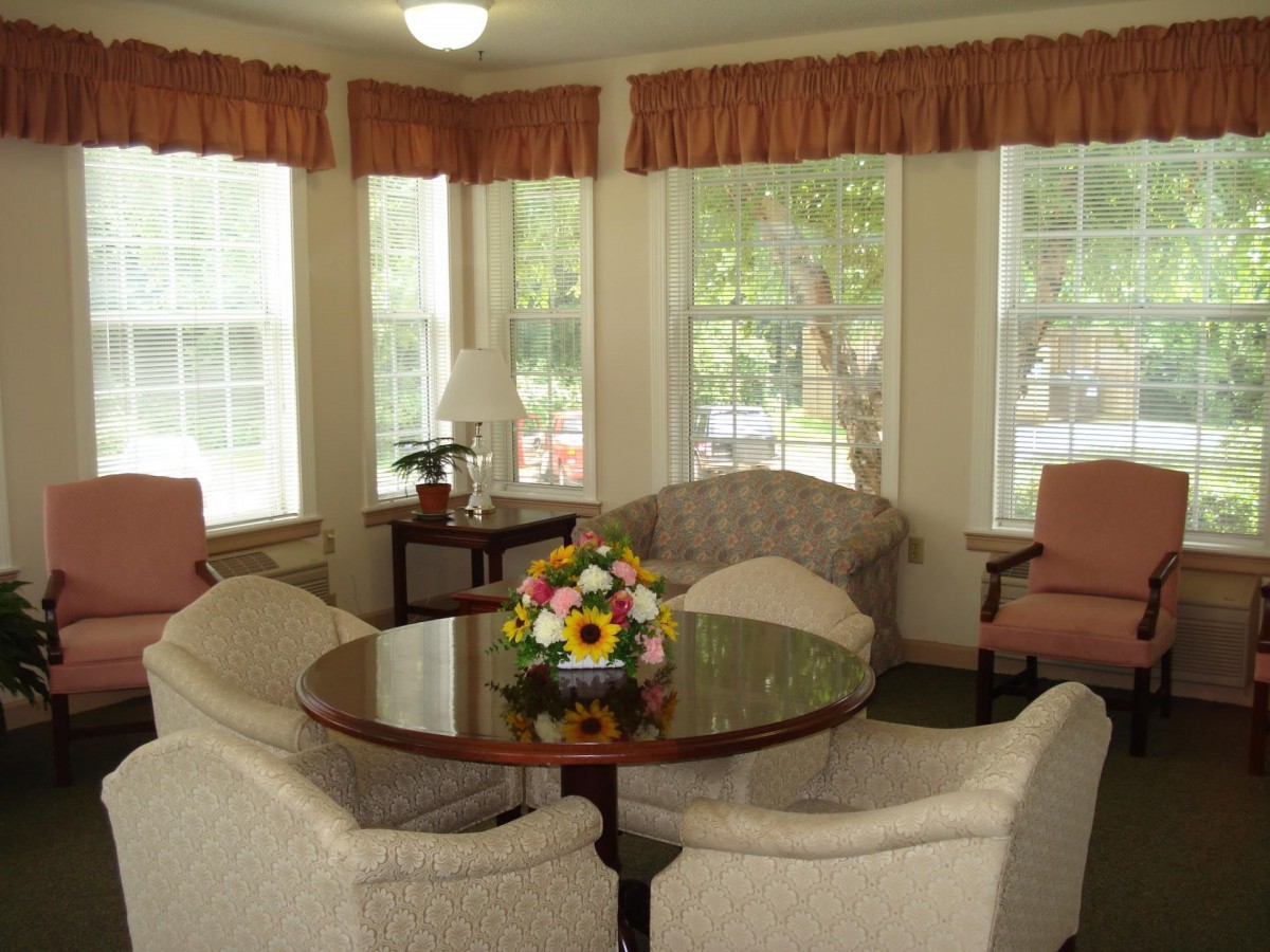 A cozy living room with a round table and comfortable chairs surrounded by large windows with light curtains.