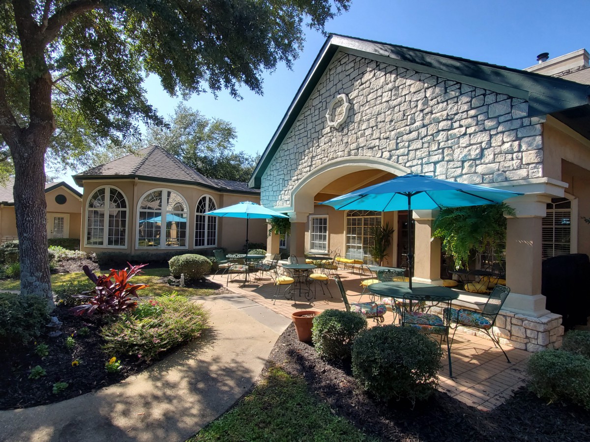 A charming outdoor patio area with tables and umbrellas, surrounded by lush garden landscaping and architectural features under a clear blue sky.
