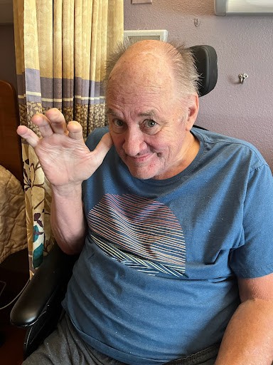 A smiling man in a wheelchair waving his hand indoors.