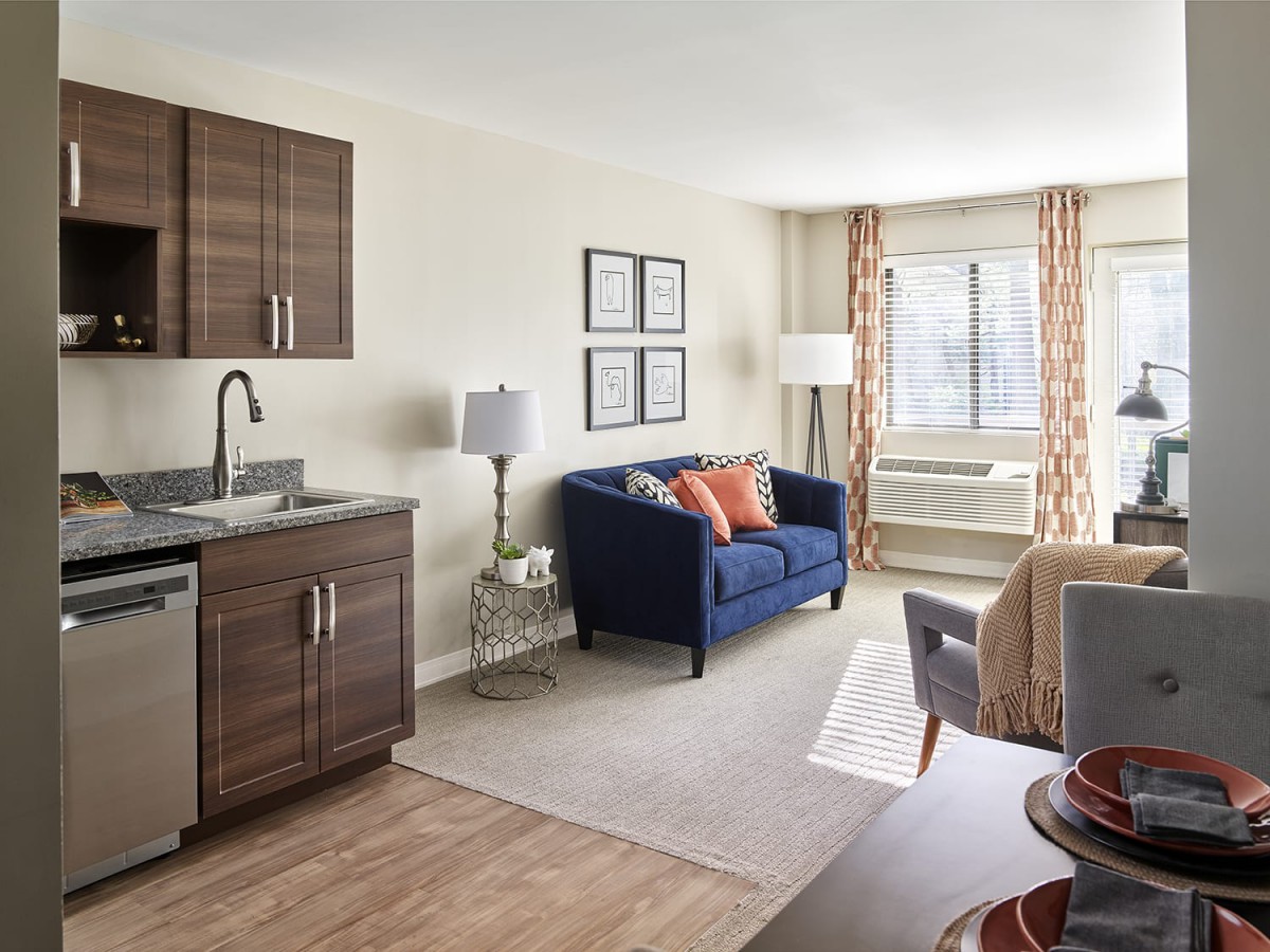 A modern apartment living area with a kitchenette, blue sofa, and large window.