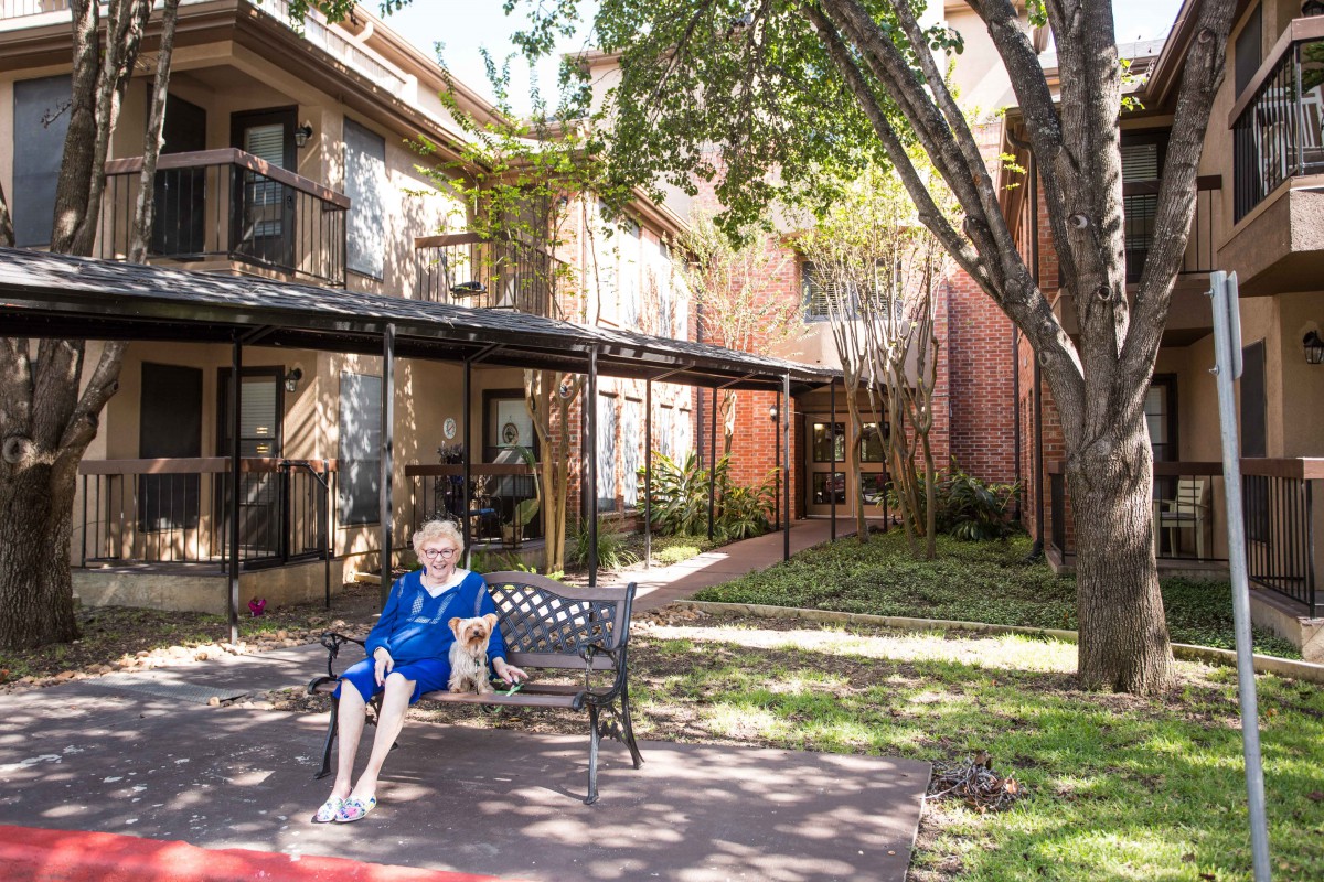 An elderly woman sits on a bench in a garden in front of a building.