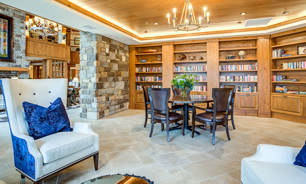 A modern library interior with wooden bookshelves, a round table surrounded by chairs, and a stone wall.