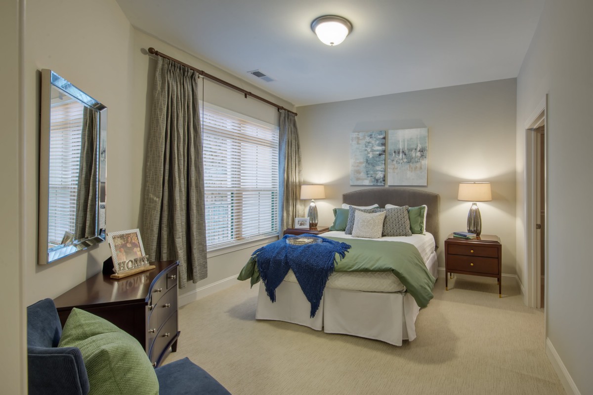 A cozy bedroom with a neatly made bed, two bedside lamps, and large windows with curtains.