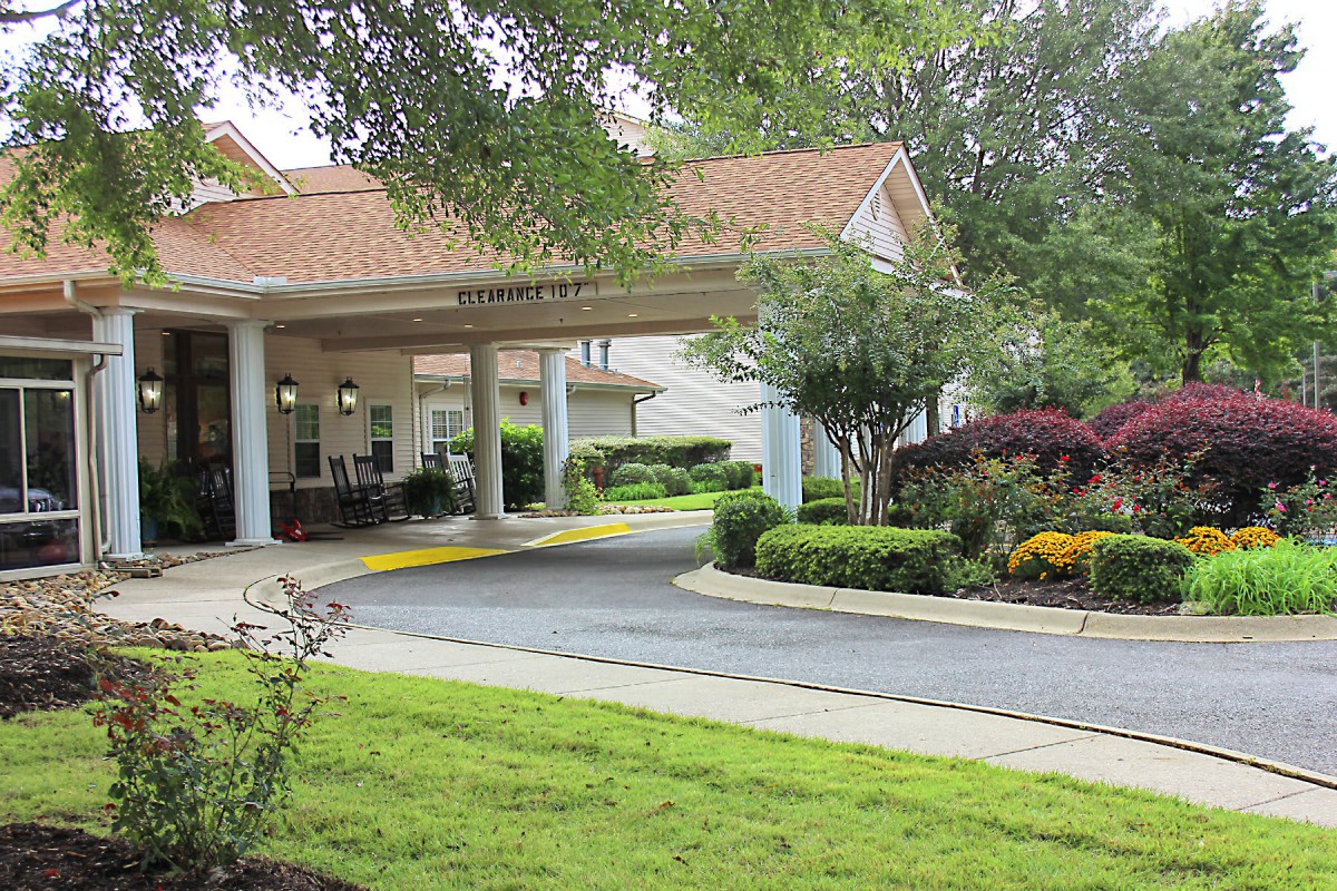 A building entrance with a landscaped driveway surrounded by trees, shrubs, and flowers.