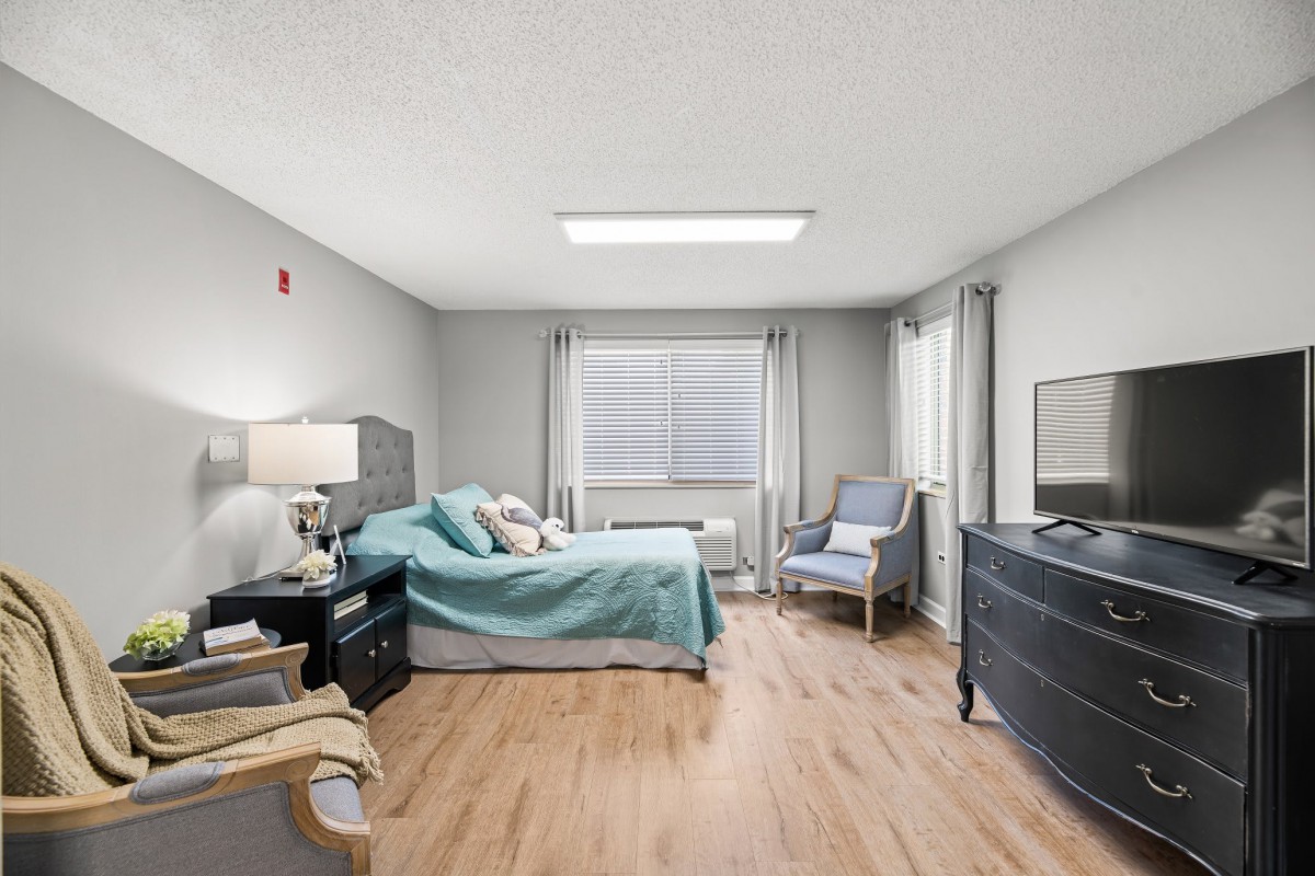 A modern and spacious bedroom with a bed, armchair, dresser, and large window.