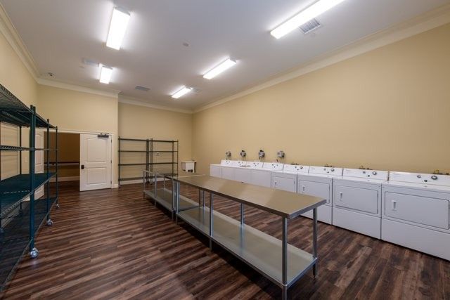A spacious laundry room with multiple washing machines and dryers, featuring wooden flooring and overhead fluorescent lighting.