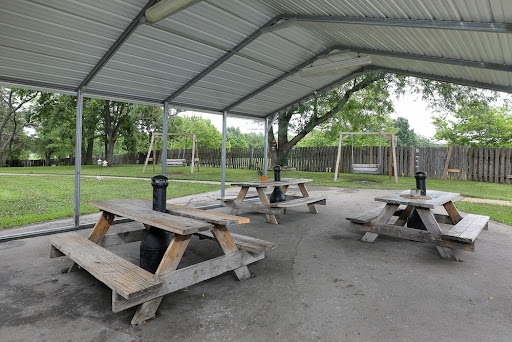 A spacious picnic area with wooden tables under a shelter, adjacent to a playground with swings.