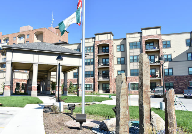 A modern residential apartment complex with a landscaped entrance and national flags flying in the foreground.