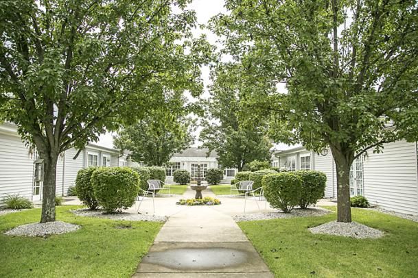 A serene outdoor garden with a central pathway, flanked by lush green trees and neatly trimmed bushes, leading to a decorative fountain.