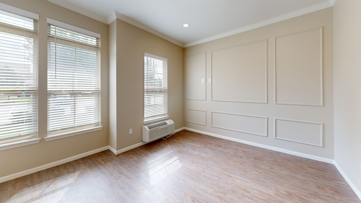 A well-lit empty room with hardwood flooring, beige walls, and two large windows letting in natural light.