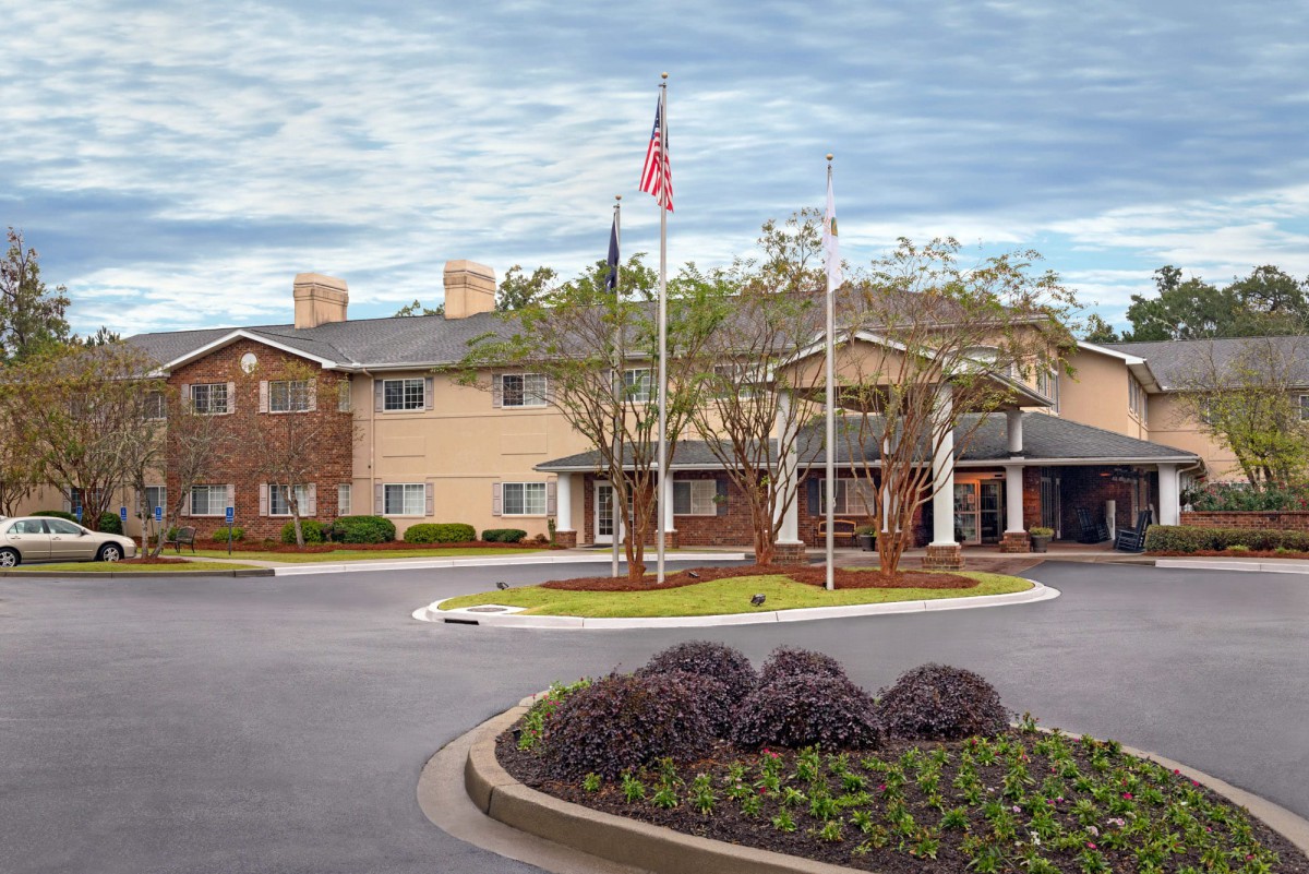 A view of a building with an entrance and flags, surrounded by landscaped greenery under a cloudy sky.