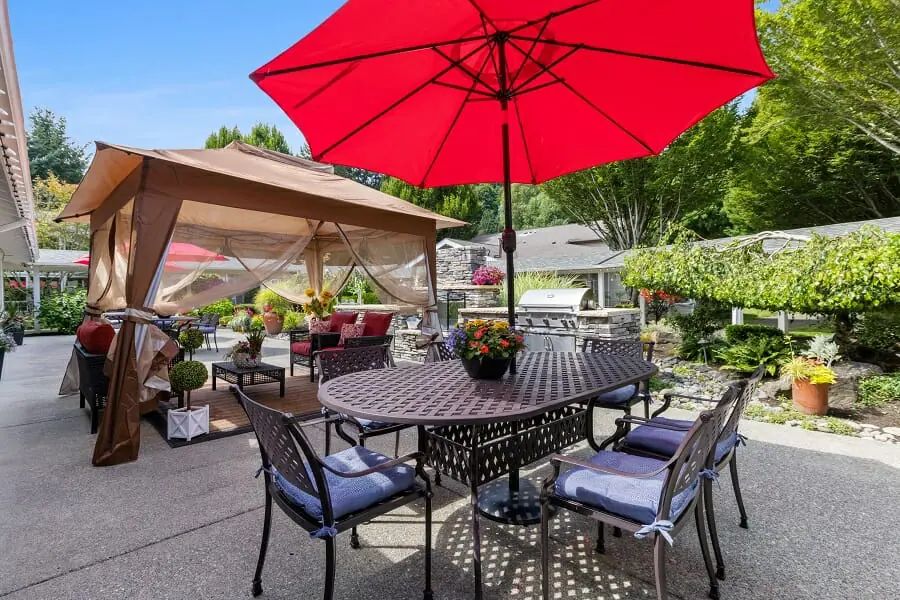 A cozy outdoor patio setup with a red umbrella shading a table and chairs, surrounded by a lush garden.