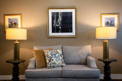 A cozy living room setup with a gray sofa, decorative pillows, and warm lighting from two lamps. A framed piece of art hangs above the sofa.