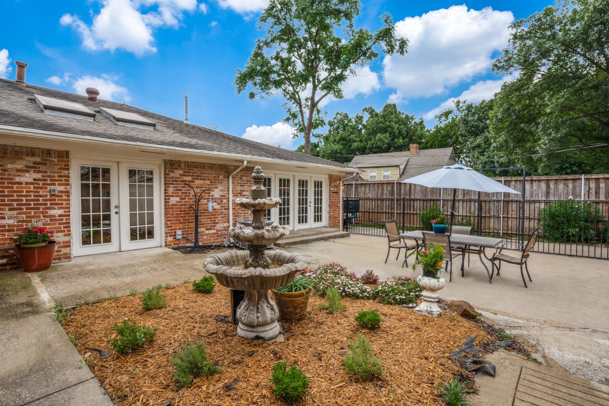 A charming backyard with a garden and fountain, featuring outdoor seating on a patio next to a brick house.