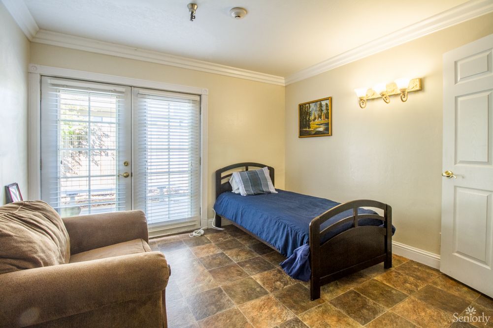A cozy bedroom featuring a single bed with blue bedding, a comfortable chair, and ample natural light from large windows.