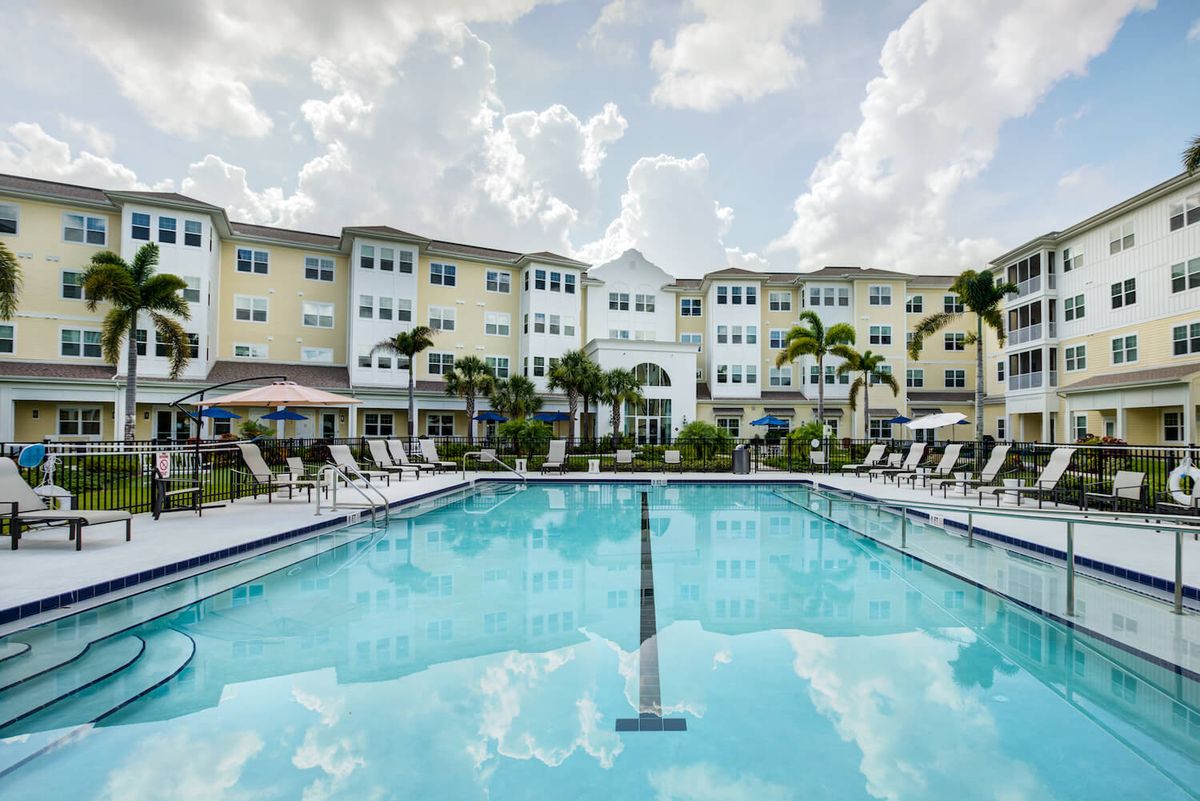 A scenic view of a swimming pool in front of a residential building surrounded by palm trees and lounge chairs under a cloudy sky.