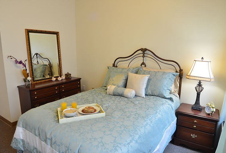 A cozy bedroom with a neatly made bed, decorative pillows, and a breakfast tray. A mirror is placed above a dresser next to the bed.