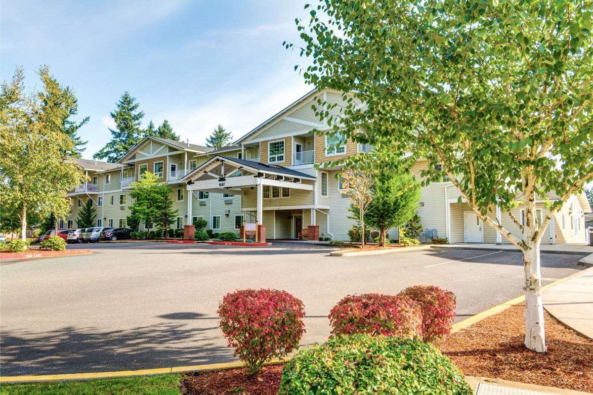 A sunny day view of a building surrounded by trees and bushes with a driveway in front.