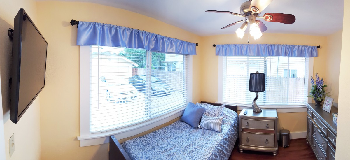 A cozy bedroom with a single bed, blue curtains, and yellow walls, illuminated by natural light from large windows.