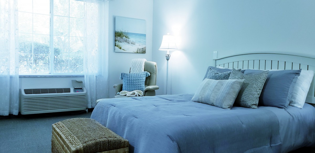 A cozy bedroom with a blue color scheme, featuring a comfortable bed, decorative pillows, an armchair, and natural light streaming through the window.