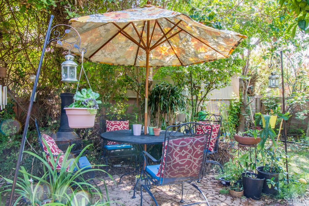 A cozy garden patio with a table, chairs, and a large umbrella surrounded by lush greenery.