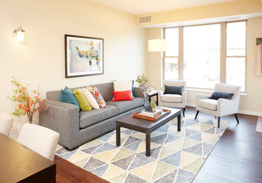 A bright and cozy living room with a gray sofa, colorful pillows, a geometric rug, and natural light.