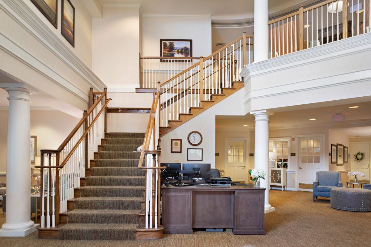 A well-lit interior space featuring a staircase with wooden railings, a reception desk, and decorative columns.