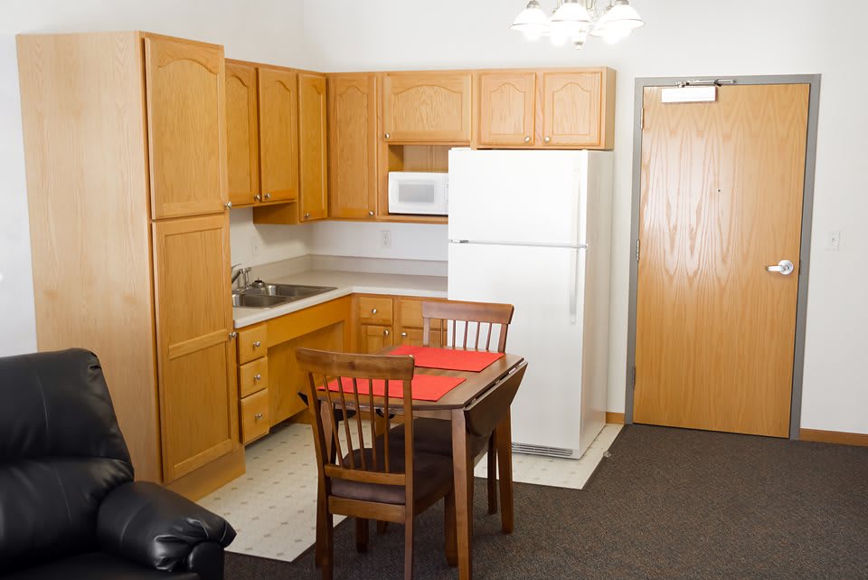 A compact kitchen in an apartment featuring wooden cabinets, a white refrigerator, a microwave, and a small dining table with chairs.