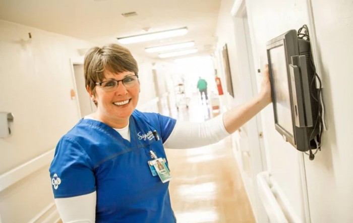 A smiling nurse in a blue uniform stands in a hospital corridor.