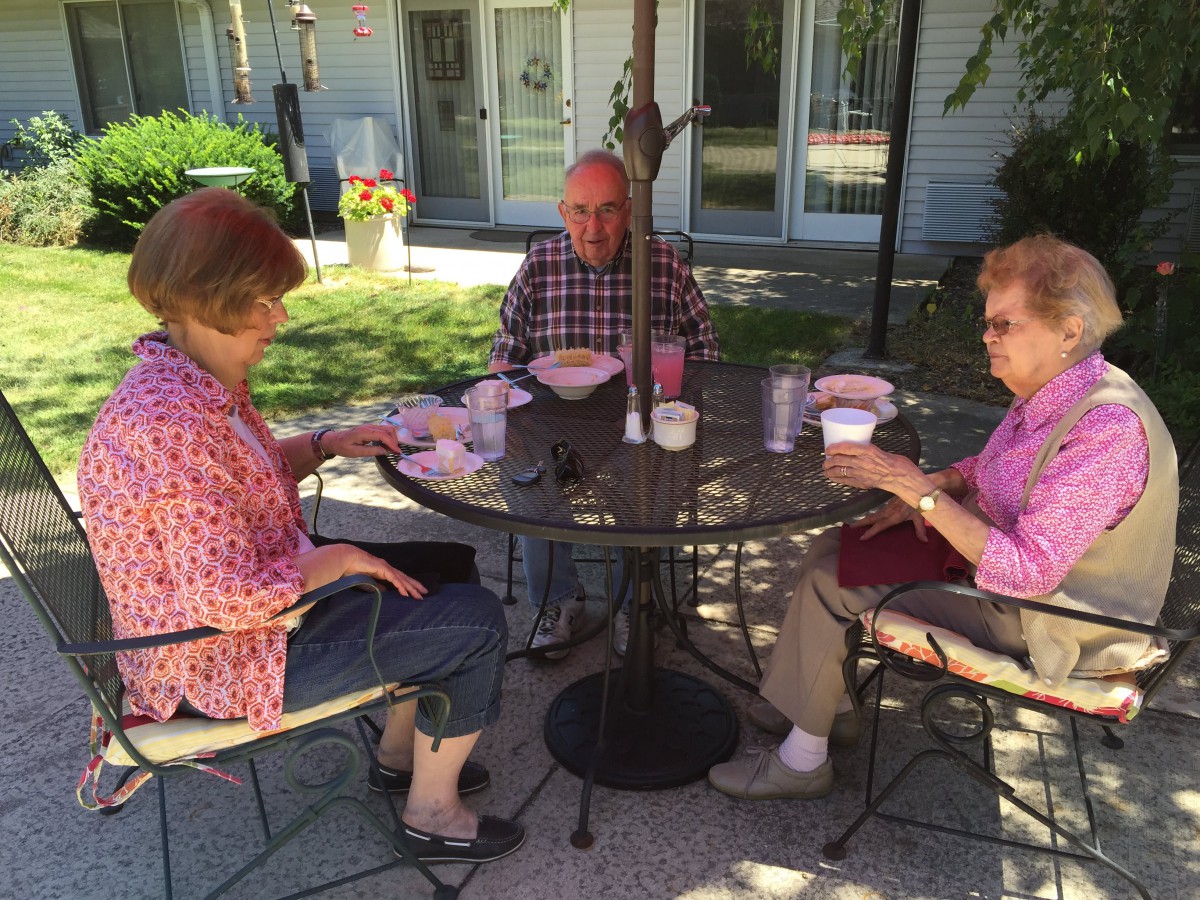 A group of three people enjoying a meal together at an outdoor table in a garden setting.