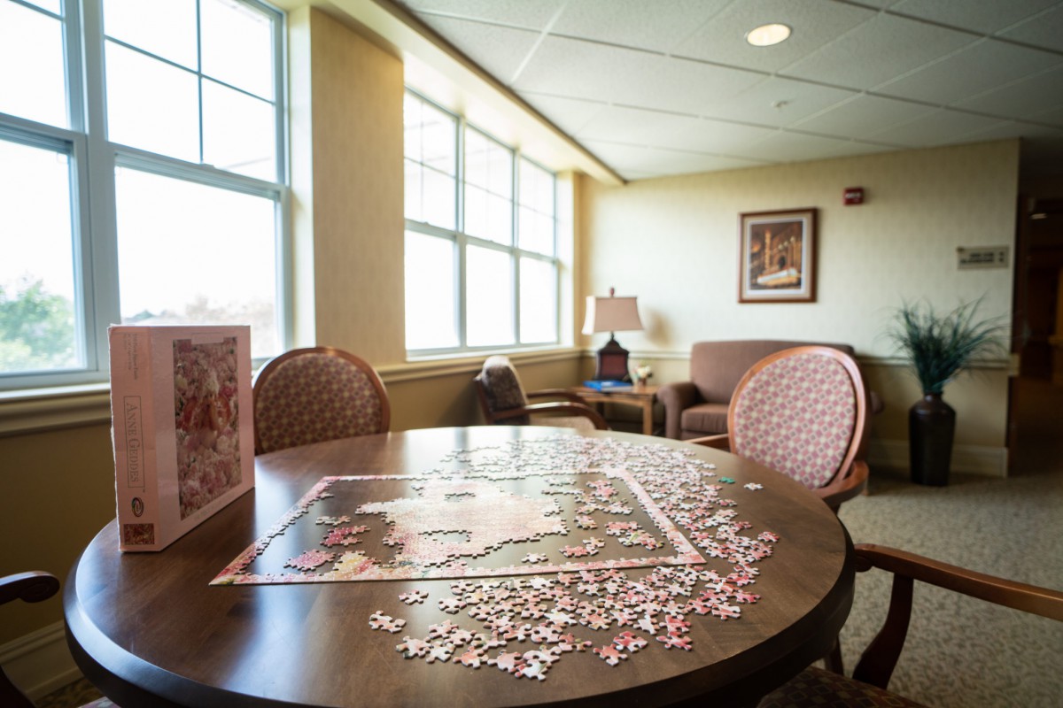 A cozy lounge area with a puzzle in progress on the table, surrounded by chairs and bathed in natural light from large windows.