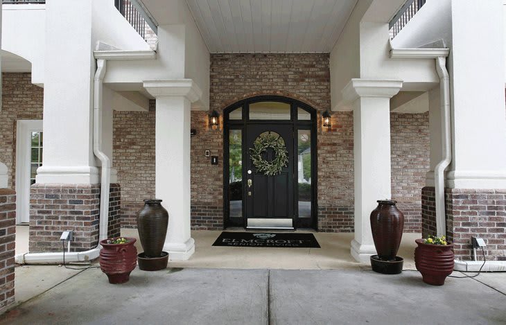 A welcoming home entrance with a black front door, surrounded by brick walls and white columns, adorned with decorative vases and a wreath.