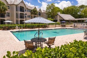 A serene outdoor swimming pool area with patio furniture and an umbrella, surrounded by residential apartment buildings and lush greenery.