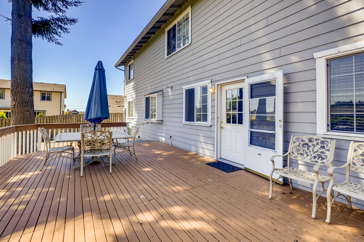 A sunny outdoor patio with a table and chairs under an umbrella on a wooden deck attached to a house.