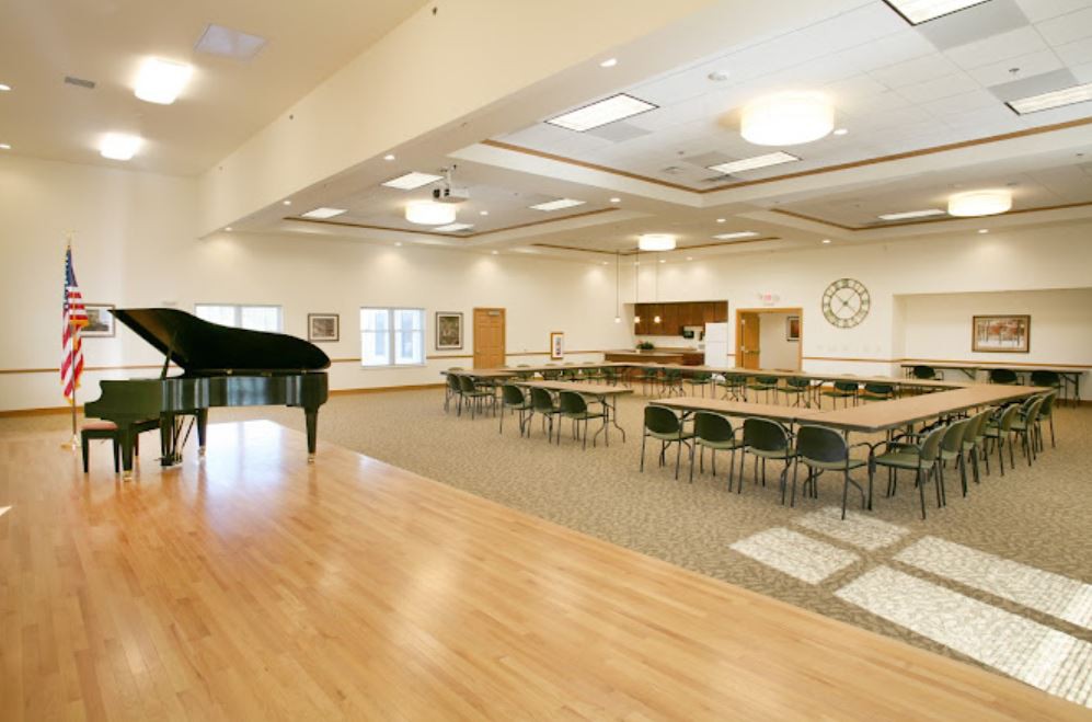 A spacious event hall with a piano on stage and several tables and chairs arranged for a meeting or gathering.